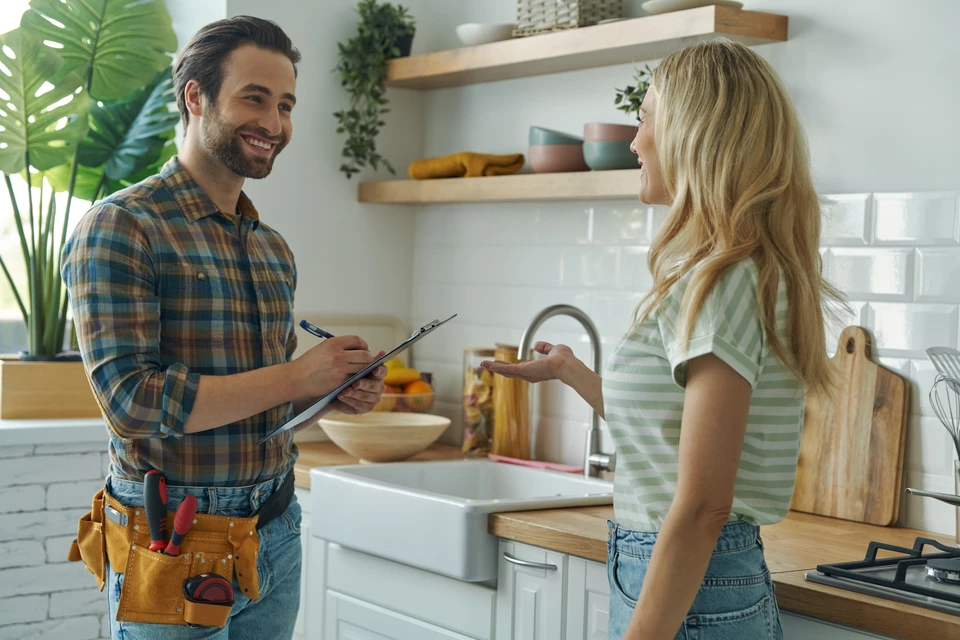 Artisan plombier-chauffagiste en tenue professionnelle discutant avec un client dans un intérieur résidentiel (salon ou cuisine), montrant plans ou devis en main, tous deux examinant attentivement le document. L'atmosphère doit véhiculer la confiance, l'écoute et le conseil technique personnalisé. Arrière-plan montrant un intérieur domestique chaleureux et accueillant.