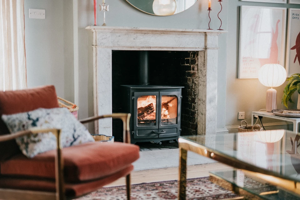 Photographie d'intérieur résidentiel montrant un salon chaleureux avec un poêle à bois ou à pellet allumé, diffusant une flamme visible. La pièce révèle une ambiance conviviale et douillet, avec de la lumière chaude provenant du feu. On peut voir des éléments qui renforcent l'atmosphère d'intimité et de confort (comme un canapé, des coussins, etc.). L'image doit capturer l'essence du bien-être et du rassemblement autour d'une source de chaleur authentique.